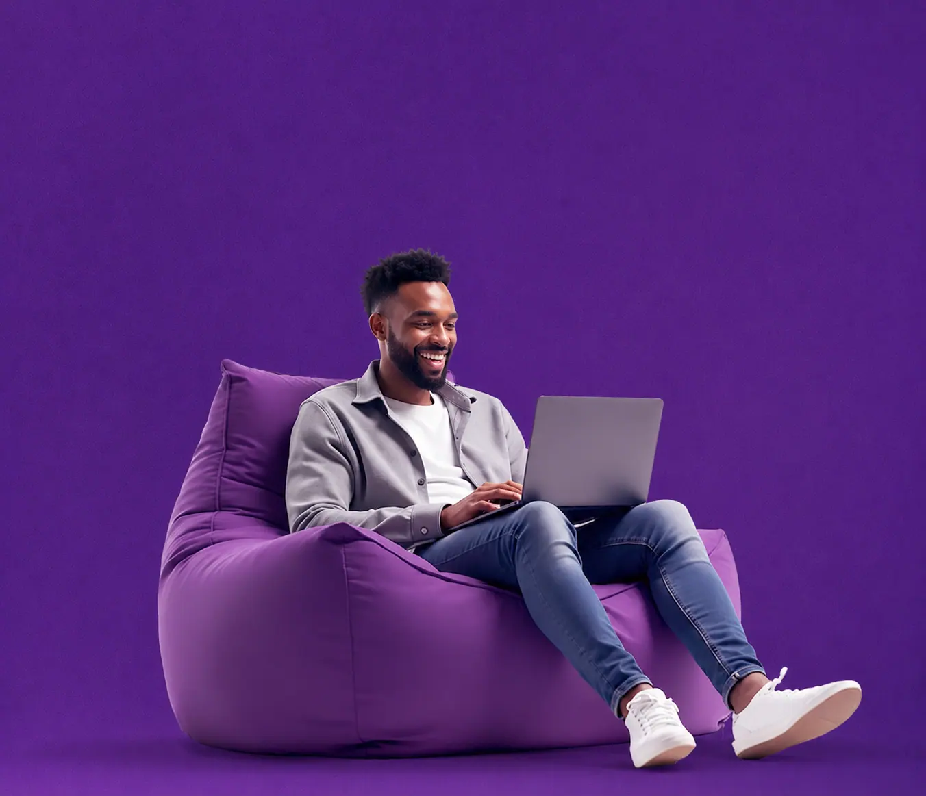 Homem sorridente trabalha relaxado em um puff roxo com o notebook no colo